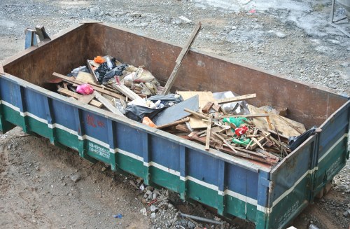 A large, open steel skip situated on a gravel and dirt surface, filled with a mixture of construction and renovation waste including wooden planks, broken panels, plastic bags, and various discarded packaging materials. The wooden planks are of different lengths and widths, some showing signs of wear and splintering. The plastic bags are black and partially torn, with debris spilling out. Surrounding the skip, the ground is uneven with loose gravel and small stones, indicating an outdoor construction or renovation site within the vicinity of Knightsbridge. Visible in the background are patches of dirt and gravel, with no other structures or items present. The skip is used by Commercial Waste Knightsbridge for waste collection and disposal related to building or renovation projects, reflecting typical waste management activities in the local area.