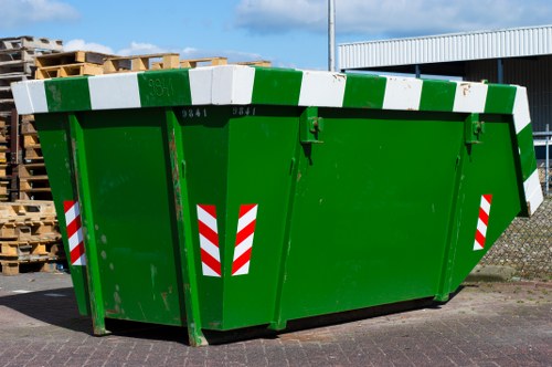 A large green commercial waste skip with red and white reflective warning stripes positioned on a paved surface outside a building, likely used for rubbish removal services in Knightsbridge. The skip has a sturdy, flat, metal lid on top, painted in alternating green and white stripes, with visible hinge mechanisms along one side. It features a smooth, glossy finish typical of industrial waste containers, and its sides are reinforced with vertical metal supports for durability. Surrounding the skip, there are wooden pallets stacked to the left, situated against a backdrop of a partly cloudy blue sky and an adjacent warehouse or industrial building with a white corrugated metal roof, contributing to the utilitarian environment typical of commercial waste collection sites. The scene captures the functional nature of rubbish disposal equipment often managed by companies like Commercial Waste Knightsbridge, serving local businesses and enterprises in the Knightsbridge area, conveniently positioned near the postcode inferred from the webpage title and URL.