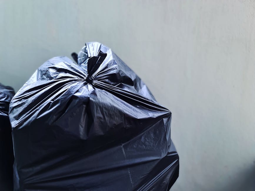 Close-up of tightly tied black plastic rubbish bags with a glossy finish, positioned in the foreground against a plain, light-colored wall. The bags are placed on a flat surface, likely pavement or co