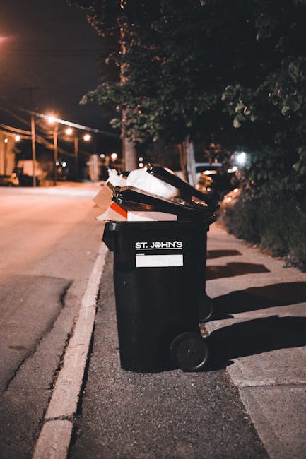 Close-up of tightly tied black plastic rubbish bags with a glossy finish, positioned in the foreground against a plain, light-colored wall. The bags are placed on a flat surface, likely pavement or co