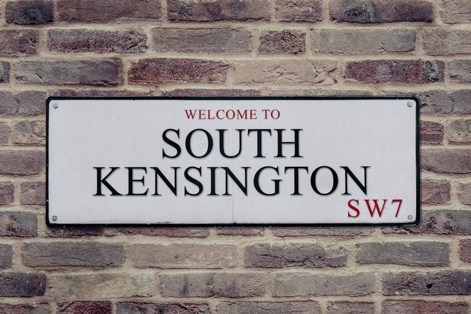 A rectangular metal street sign mounted on a brick wall, displaying the words 'WELCOME TO SOUTH KENSINGTON' in large black capital letters on a white background. Above this, in smaller red uppercase l